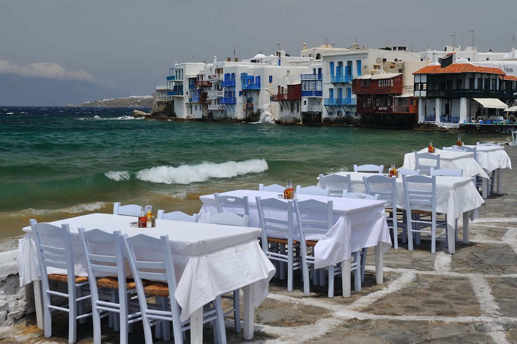 Kykladen-062009-071.jpg - Restauranttische an der Strandpromenade von Mykonos-Stadt. Der Blick ist malerisch, aber es windet hier so, dass vermutlich das Souvlaki nach einer Minute vom Tisch fliegen würde. Nicht umsonst stehen fünf Windmühlen ein Stückchen hinter mir.